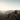 A black 2010 Lexus GX460 sits on the right side of a rocky alpine cliff above a broad mountain canyon. The wide scene features layered distant peaks, pale open sky, golden-hour light, and illuminated PRISMBEAM tail lights.