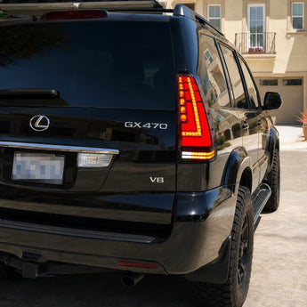 A close rear three-quarter view shows half of a black Lexus GX 470 parked in a residential driveway. The installed tail light features a red illuminated field, amber L-shaped LED strip, segmented red column, and lower amber and white sections.
