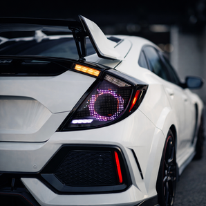 Close-up focusing on the custom digital tail light of a white 10th-gen Honda Civic hatchback. The aftermarket assembly features a smoked angular housing containing a distinctive purple pixelated LED ring. A sharp amber LED turn signal strip sits above the digital circle, with a white reverse light bar beneath it. The glowing, futuristic light pattern contrasts vividly against the vehicle's white paint and the dark, out-of-focus background, drawing full attention to the intricate electronic design.