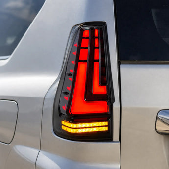This close-up shows an aftermarket LED tail light installed on a silver Lexus GX. The smoked housing contains illuminated red segmented sections, a layered L-shaped light signature, and two amber horizontal LED bars at the bottom.
